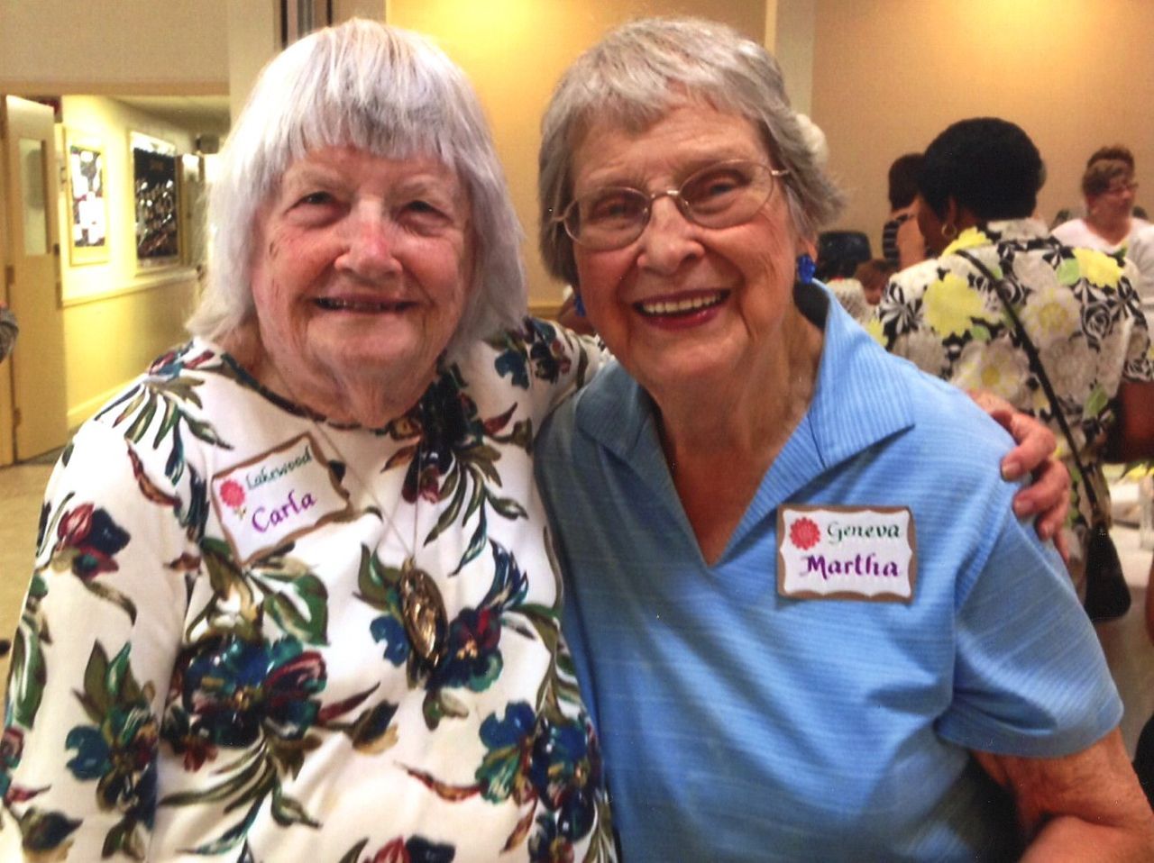 Two smiling women pose together, arms around each other. One wears a floral top, the other a blue polo. Both have name tags.