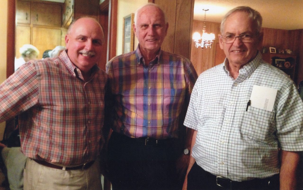Three men in button-down shirts pose indoors. The man on the left smiles, and the right man wears glasses and a name tag.