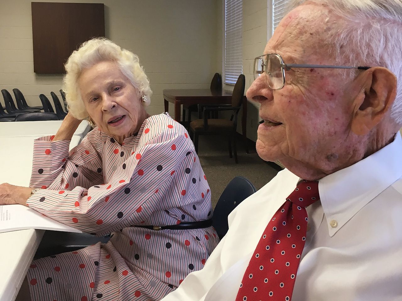An older man with glasses and a red tie speaks to a woman in a patterned dress, both seated at a table.