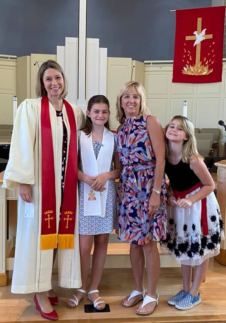 Four people in church setting; woman in robe, two girls, and another woman smiling. Red banner in background.