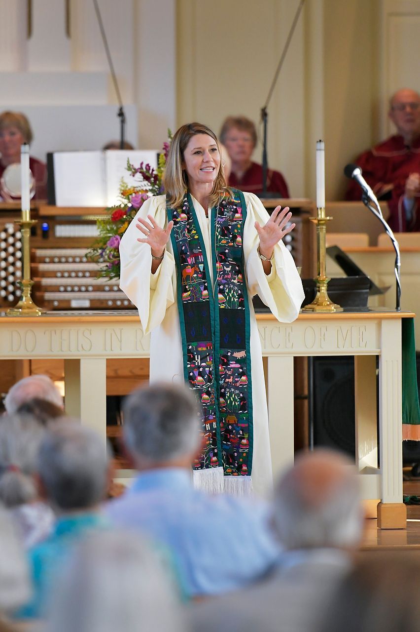 Woman preaching in a church, wearing a colorful stole, gesturing with her hands. Audience in foreground, organ in background.