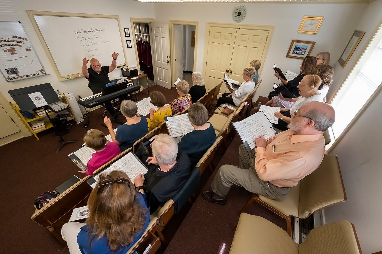 Choir practice in a small room. Conductor at keyboard directs singers holding sheet music.