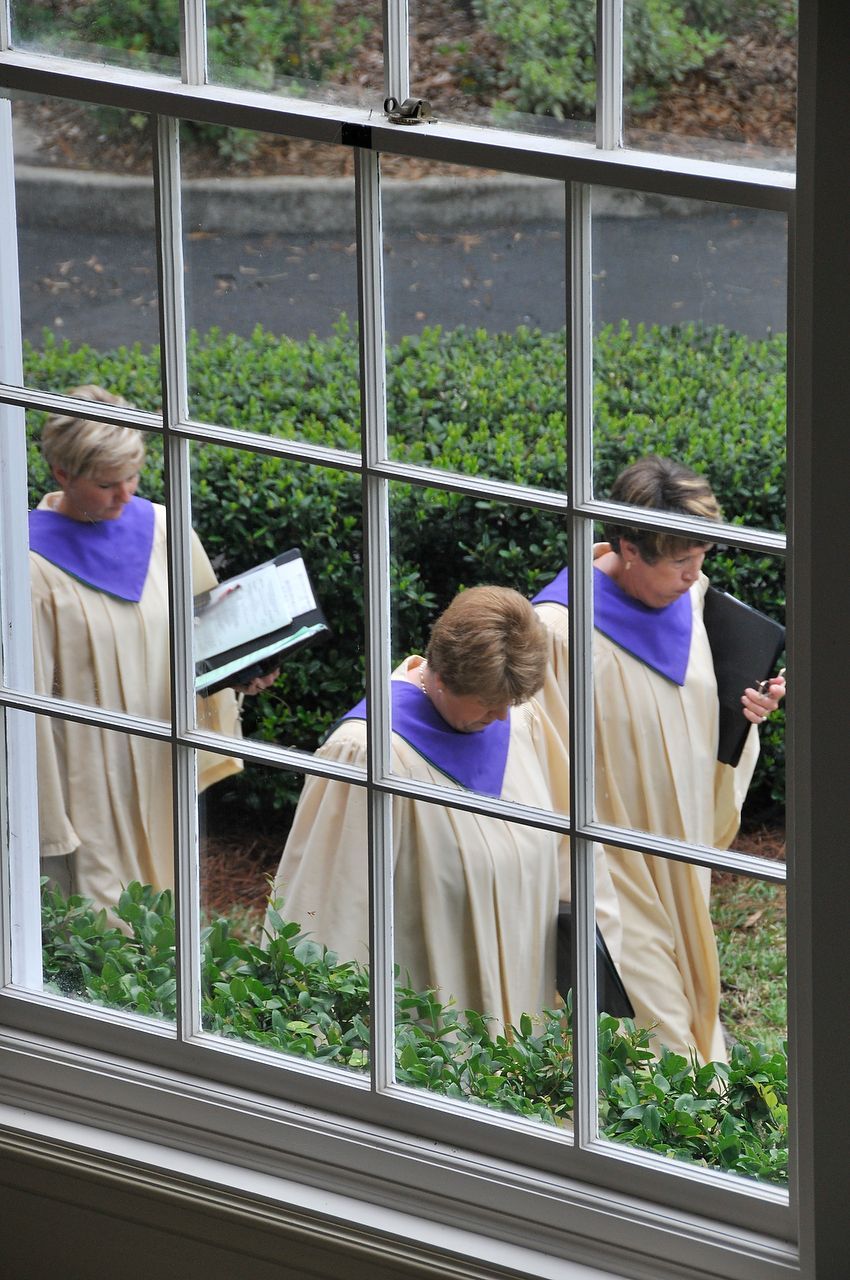 Three people in cream robes with purple collars, holding folders, walking outside past a window.