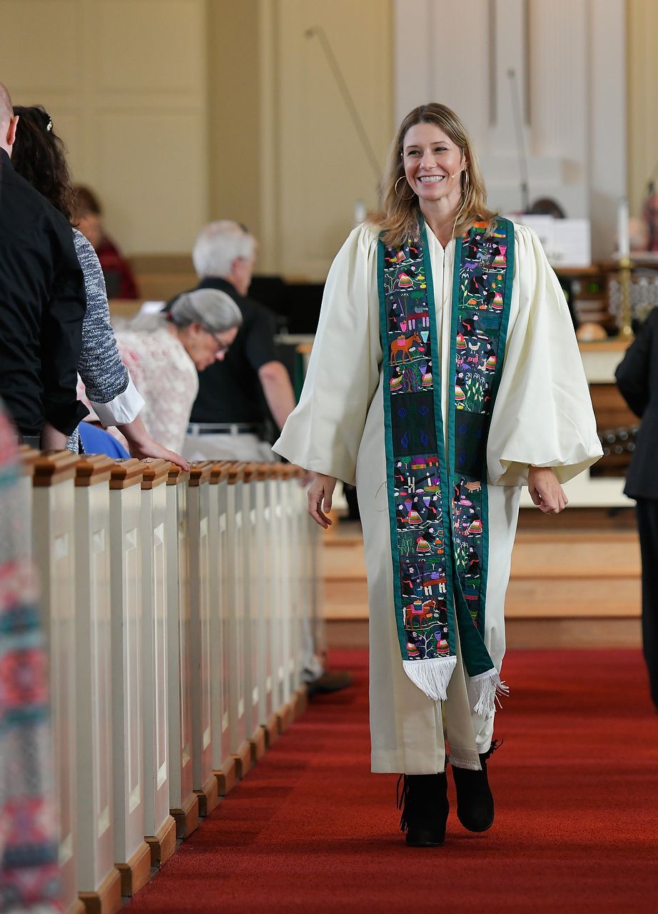 Clergy member in white robe with embroidered stole walks down a church aisle, smiling.
