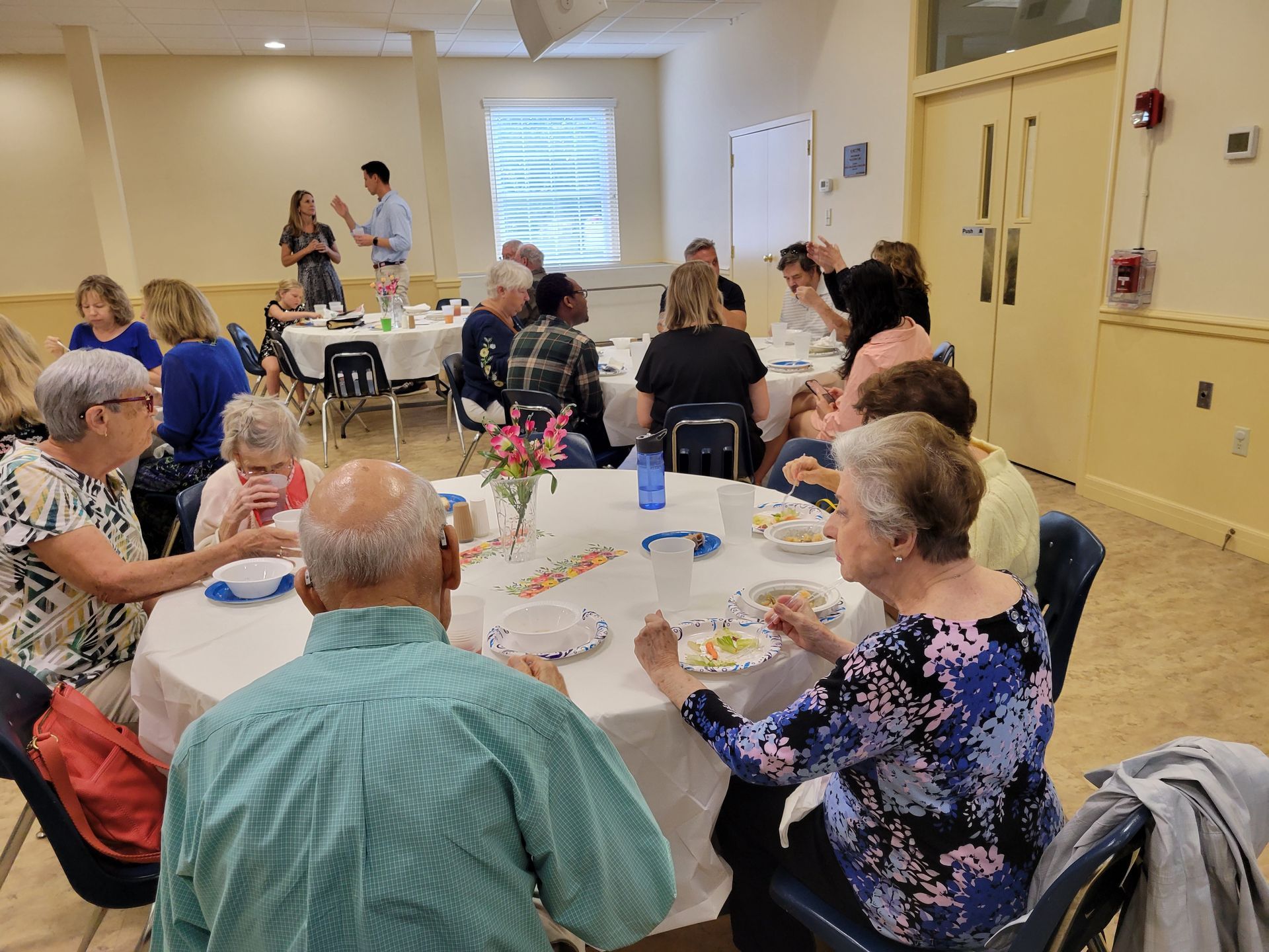 People seated at tables, eating and listening to a presentation in a brightly lit room.