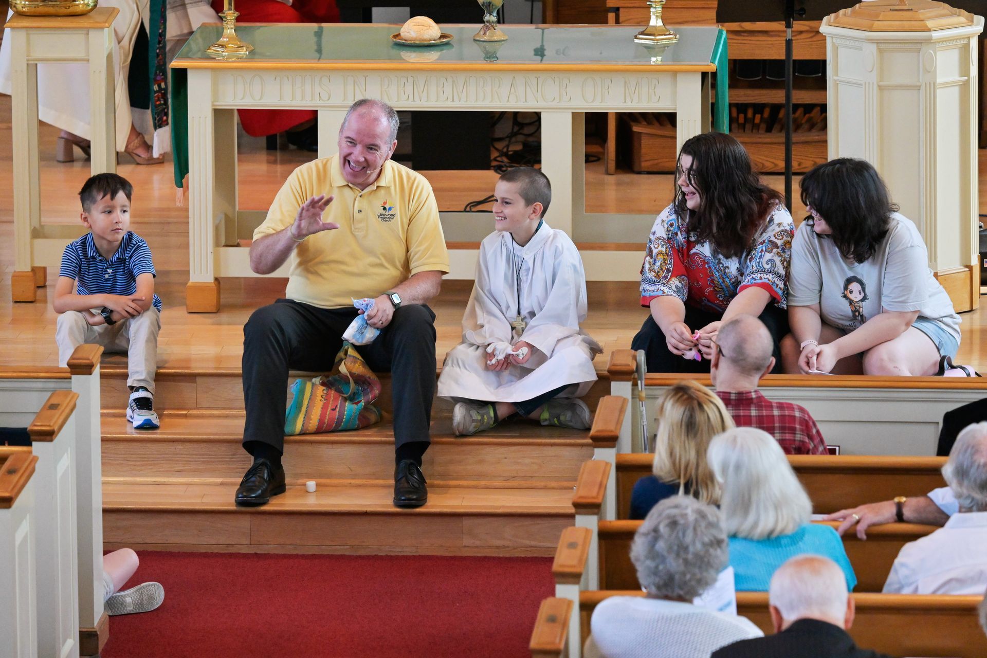 A man talking to a group of people, including children, in a church. People are sitting on pews.