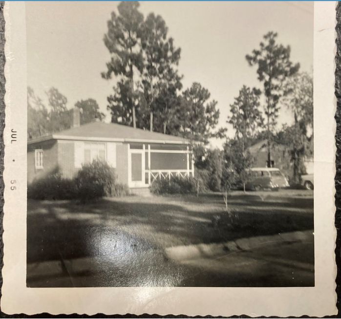 A black and white photo of a small brick house with a porch, trees, and a car in the yard.