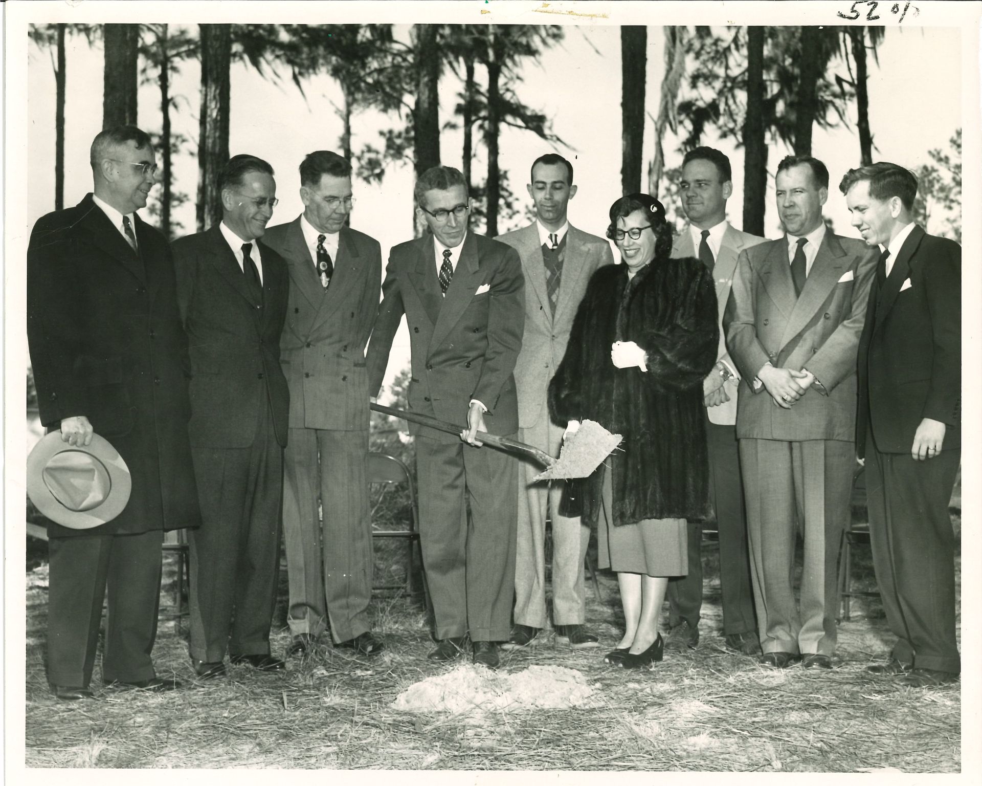 Group of men and a woman break ground with shovels, standing in a field with trees.