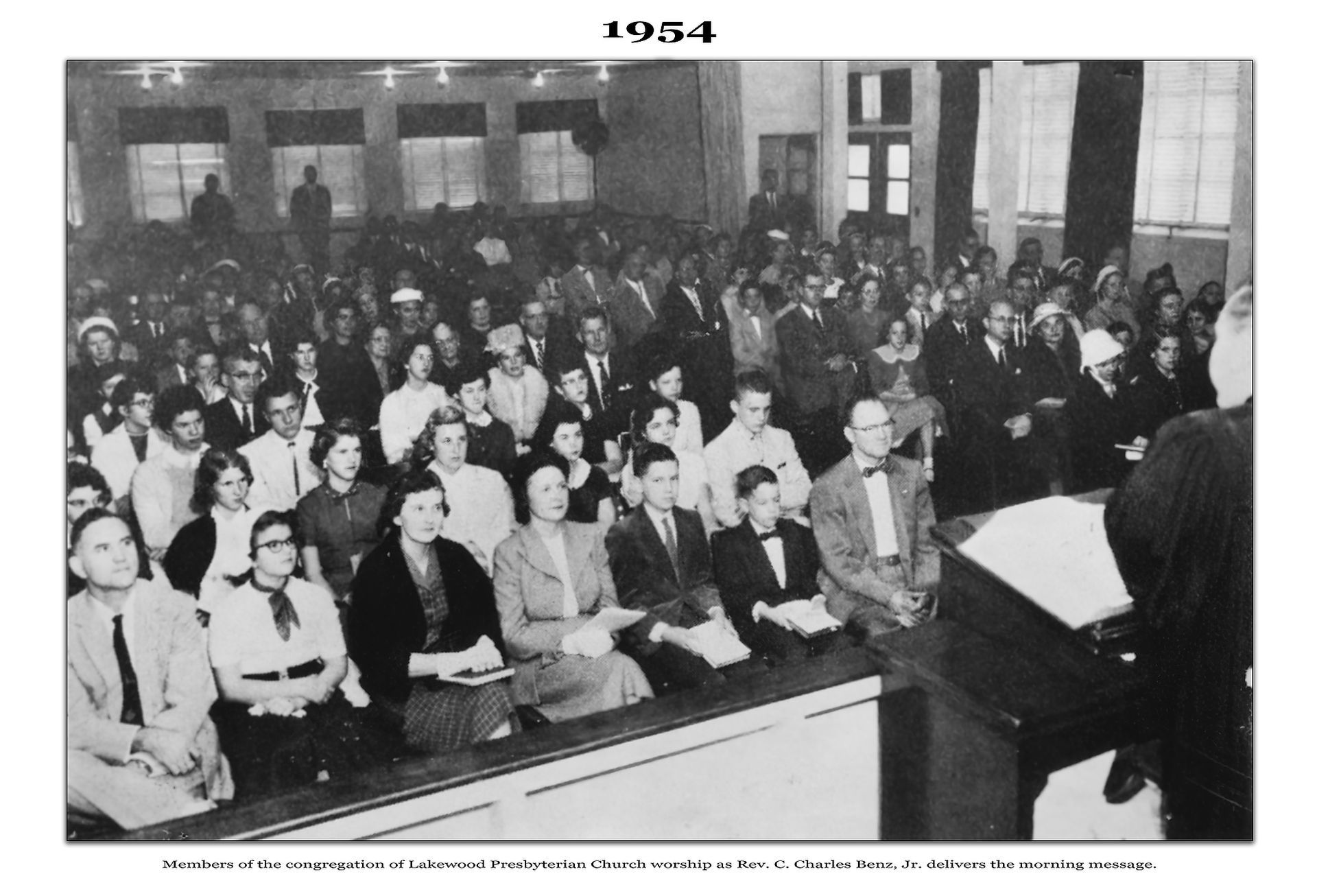 Large crowd in a room, 1954, listening to someone at a podium. Black and white photograph.