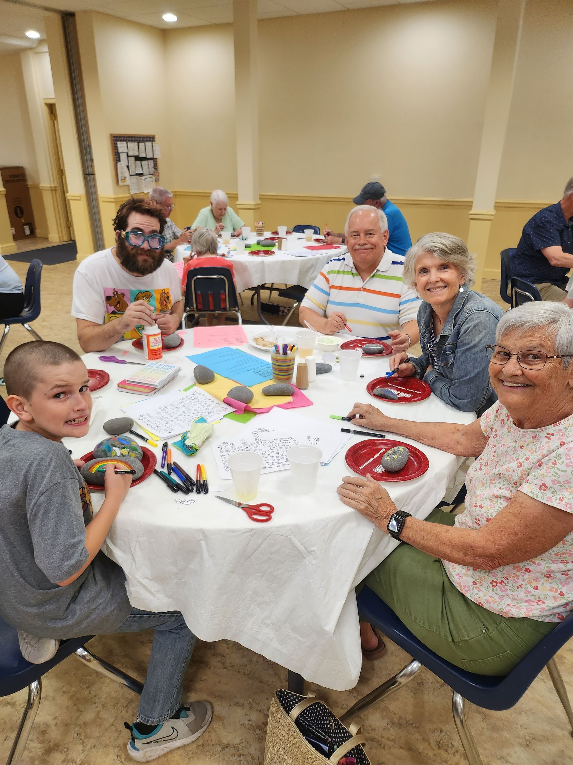 People sitting around a table, painting rocks. Colorful art supplies and smiling faces. Inside a community hall.