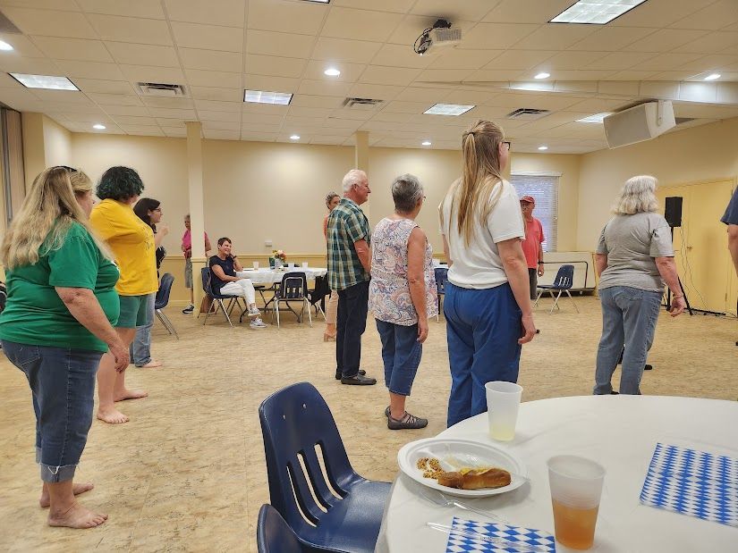 People standing in rows in a large room, some wearing casual clothes. Tables and chairs are visible.