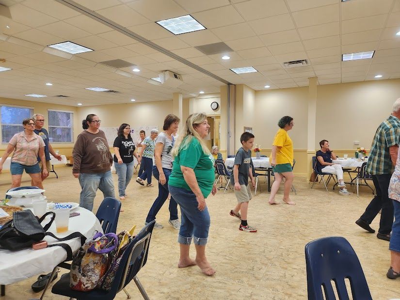 People dancing in a brightly lit room with tables and chairs. Some are wearing casual clothes.
