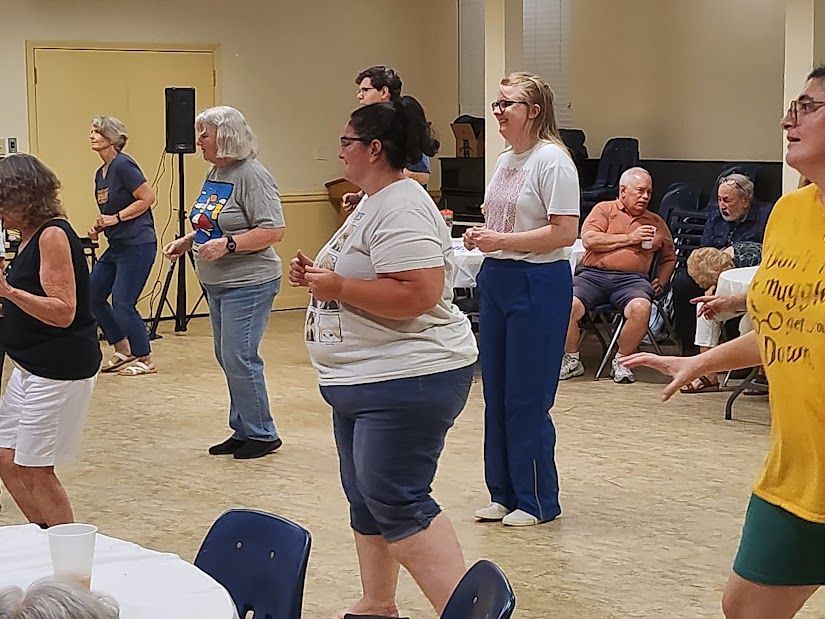 People dancing in a community hall, some watching, others near a speaker and chairs.