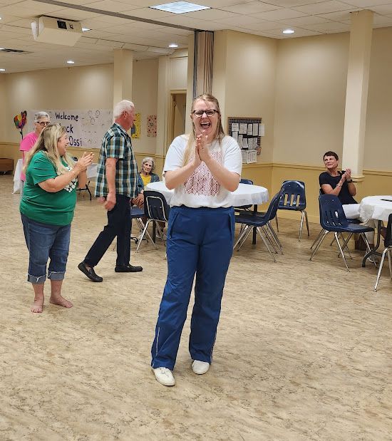Woman clapping, smiling joyfully in a room with people, tables, and a 