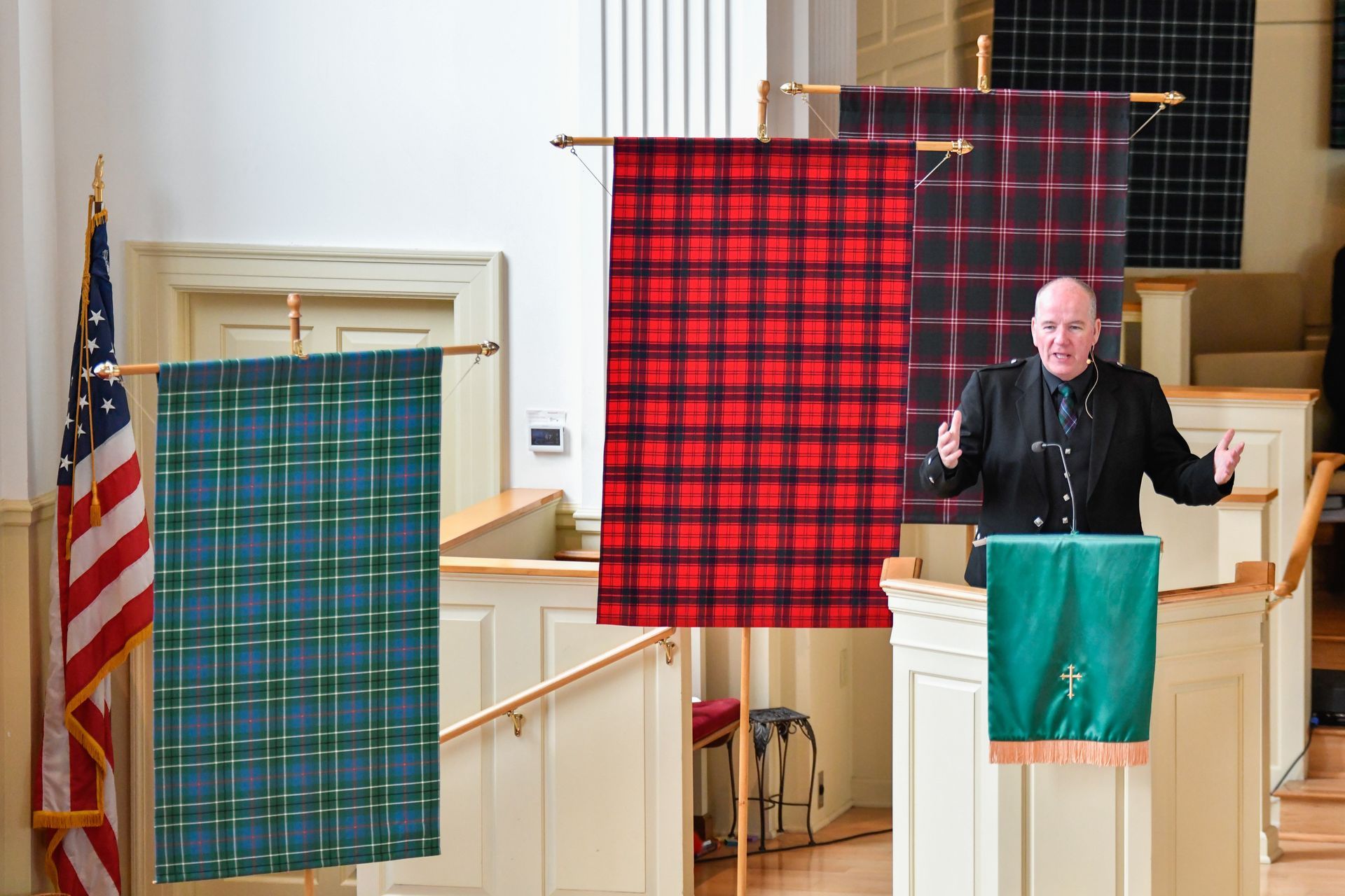 Man speaking at a podium in a church, flanked by tartan banners and an American flag.