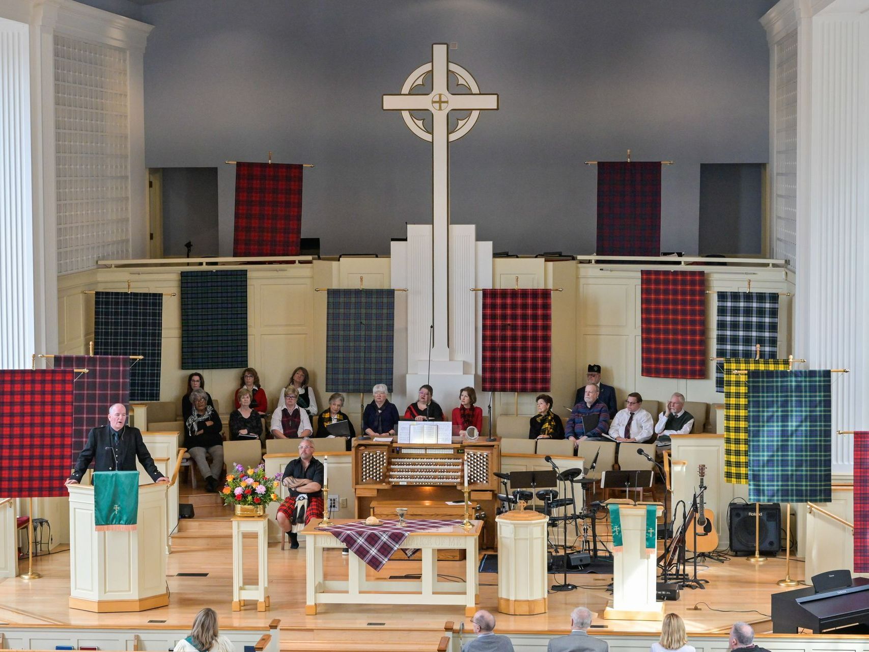 Church service, man preaching from podium; choir and congregation behind him, crosses and plaid banners.