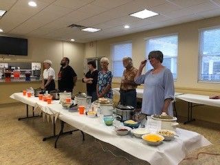 People at a chili cook-off, standing behind a table with crockpots, bowls, and toppings, inside a community room.