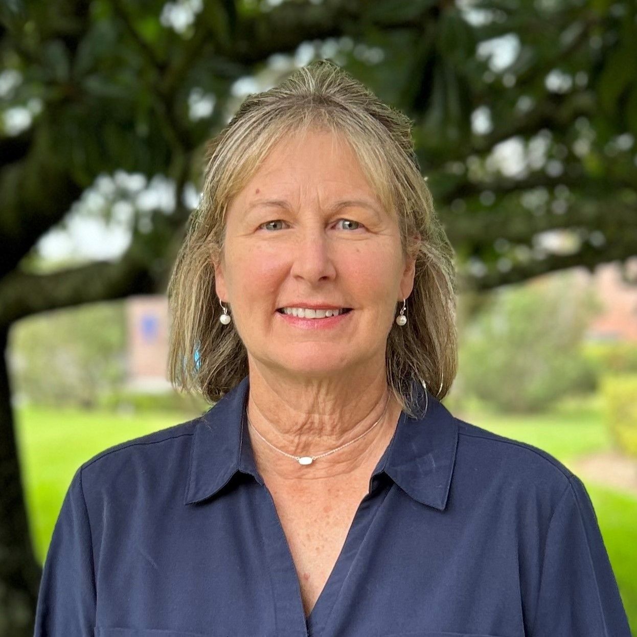 Woman smiling, wearing a dark blue shirt, with trees in the background.