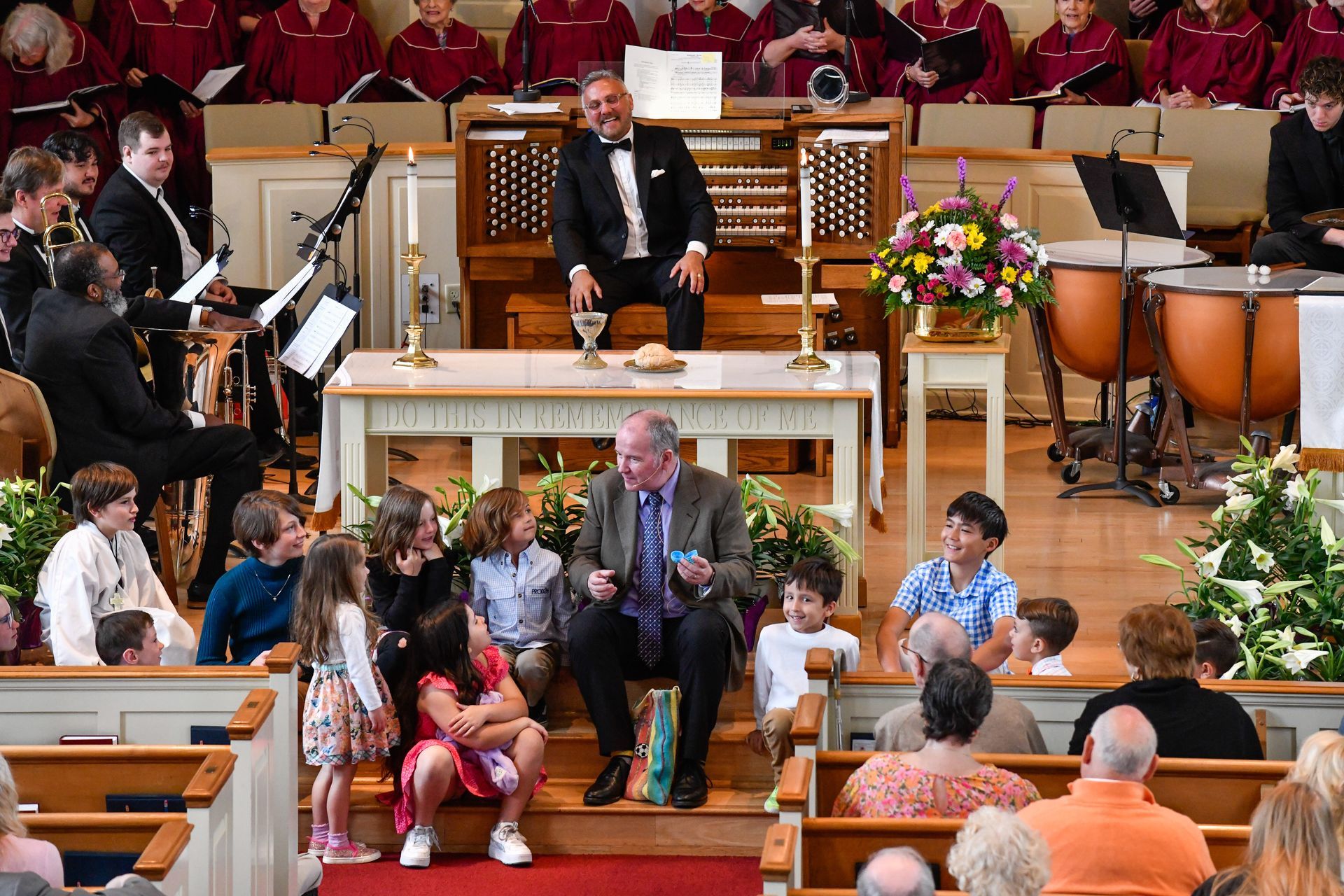 A church service with a man speaking to children, organist, and choir.