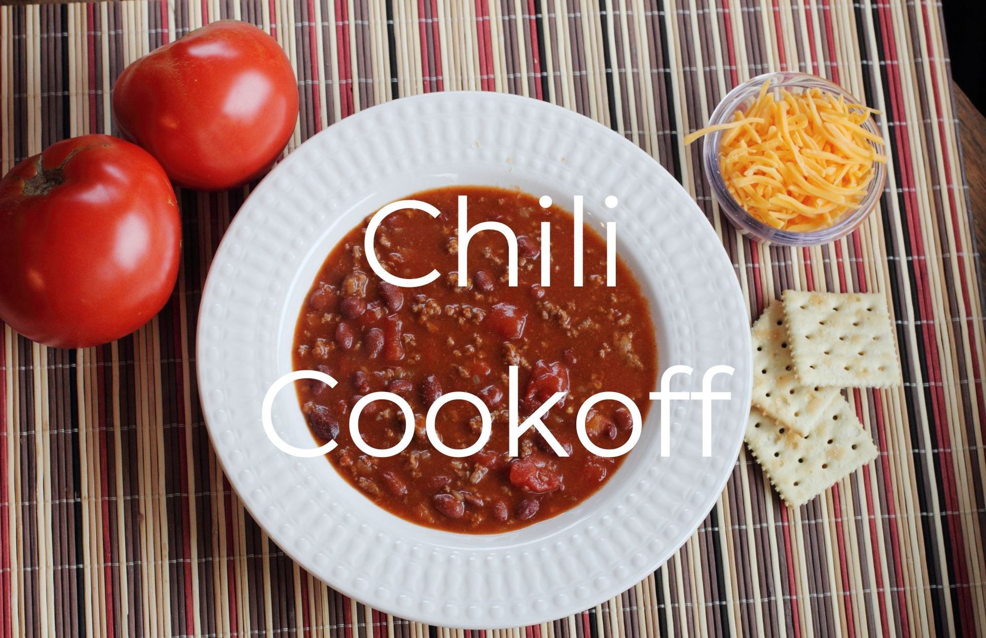Chili in white bowl, tomatoes, shredded cheese, and crackers on a mat, 
