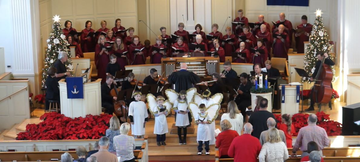 A choir and orchestra perform on a church stage decorated for Christmas. Angel children stand in front.