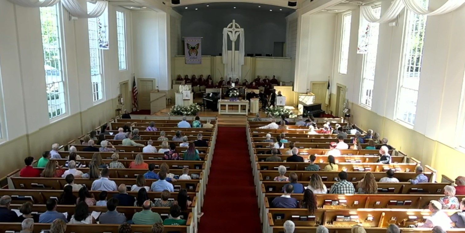 A church interior with a red carpet aisle, pews filled with people, and a choir in the background.