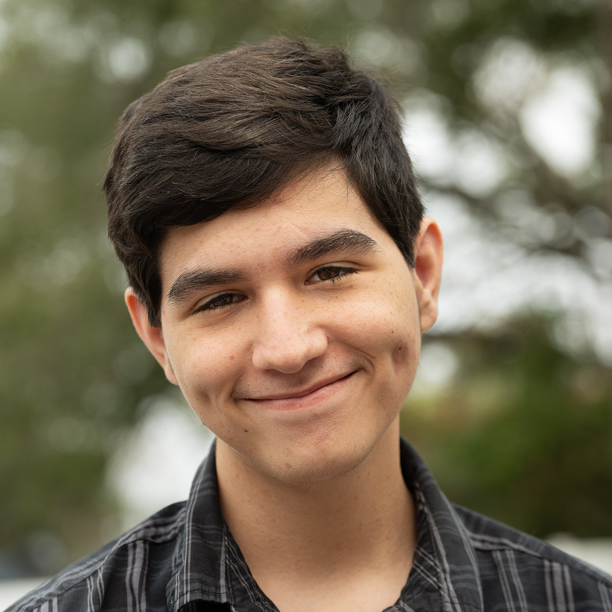 Young person with dark hair and a dimpled smile outdoors, wearing a plaid shirt.