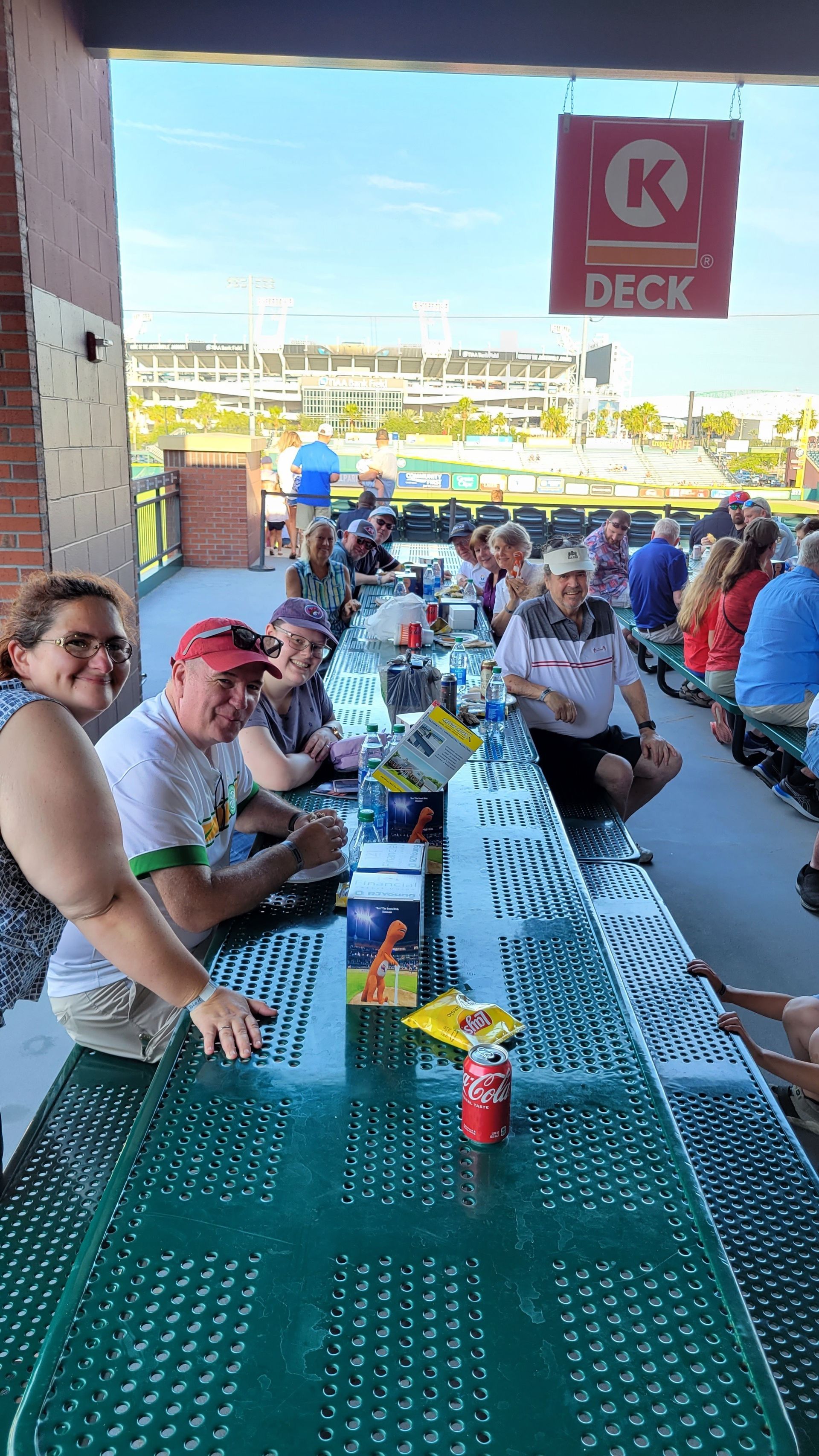 People at outdoor picnic tables under a deck; a baseball game is visible in the background.