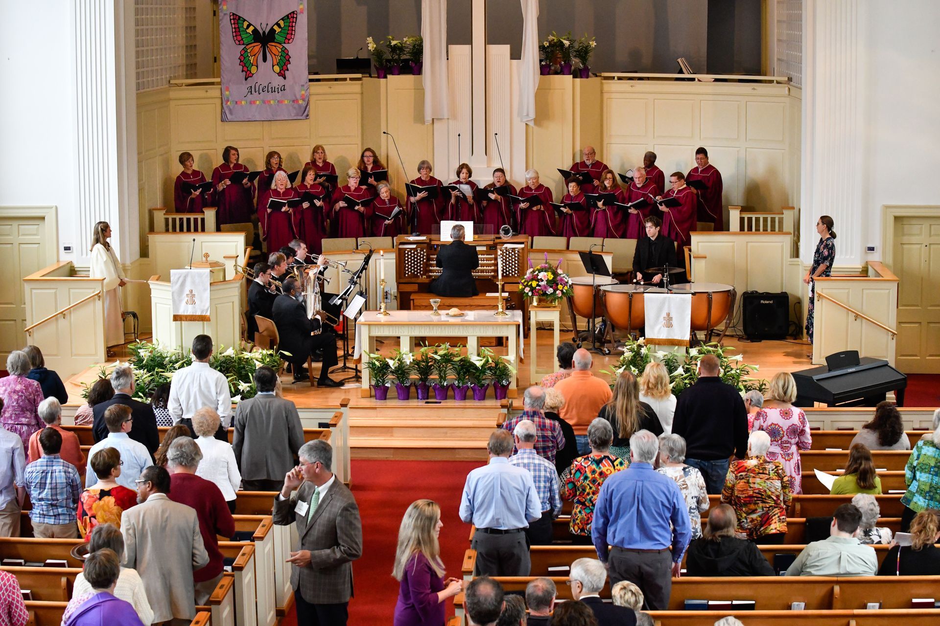 Church service with choir, orchestra, and congregation. People gather for event, flowers on altar, red carpet.