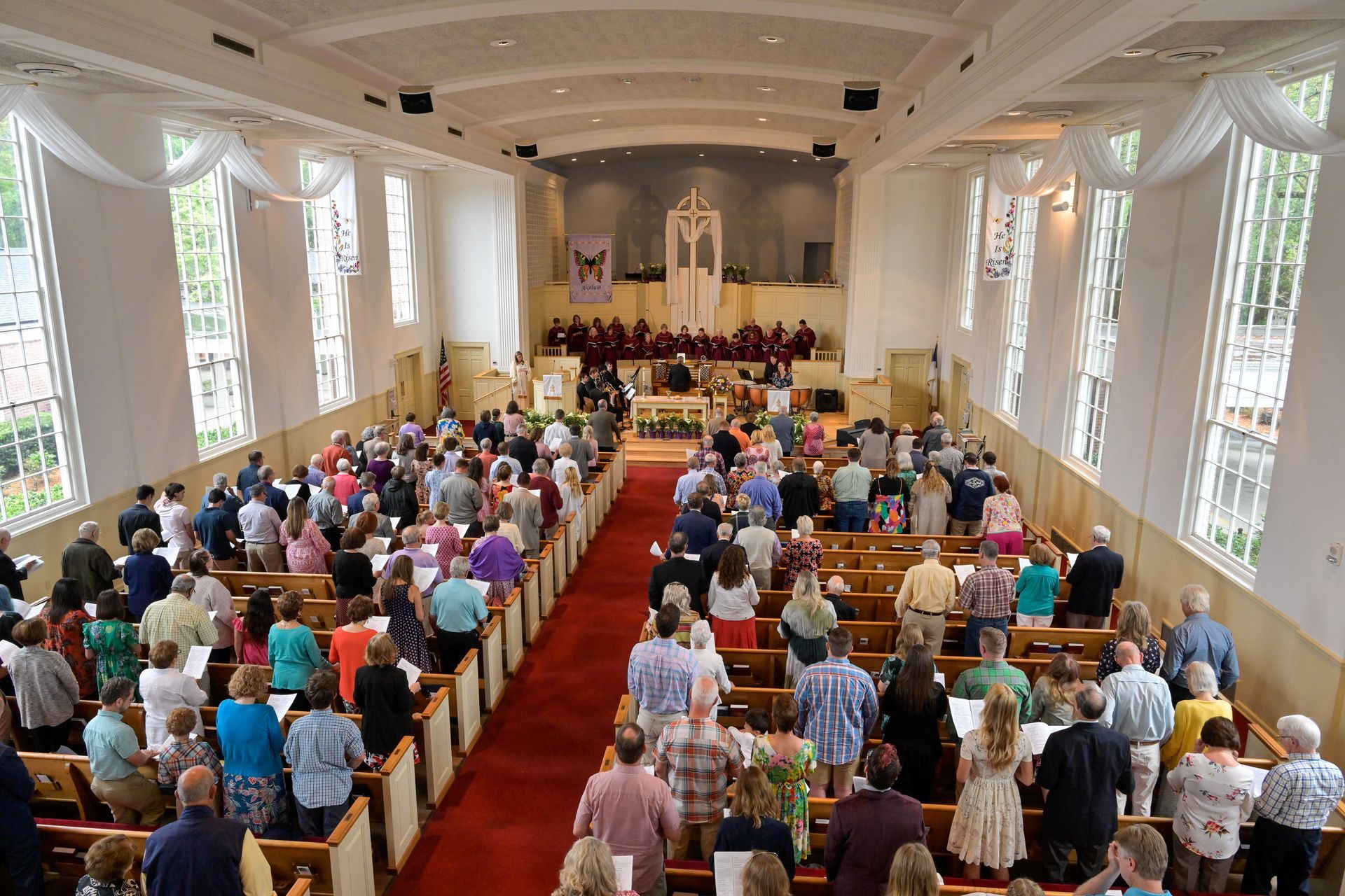 Church interior filled with people standing and facing altar, red carpet, sunlight, and large windows.