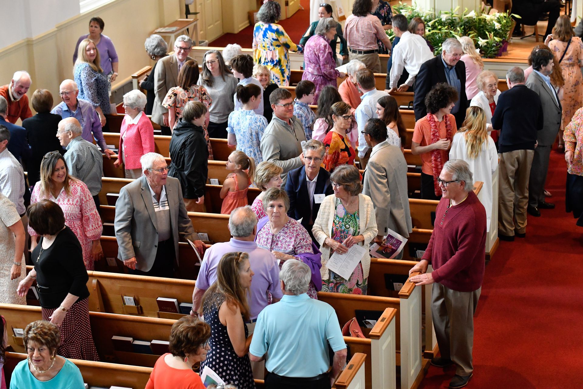 People standing and talking inside a church with wooden pews and red carpet.