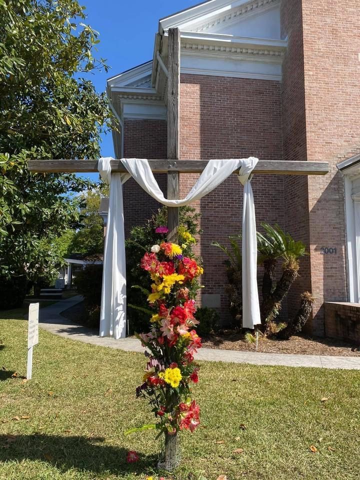 Wooden cross decorated with flowers and white fabric in front of a brick building.