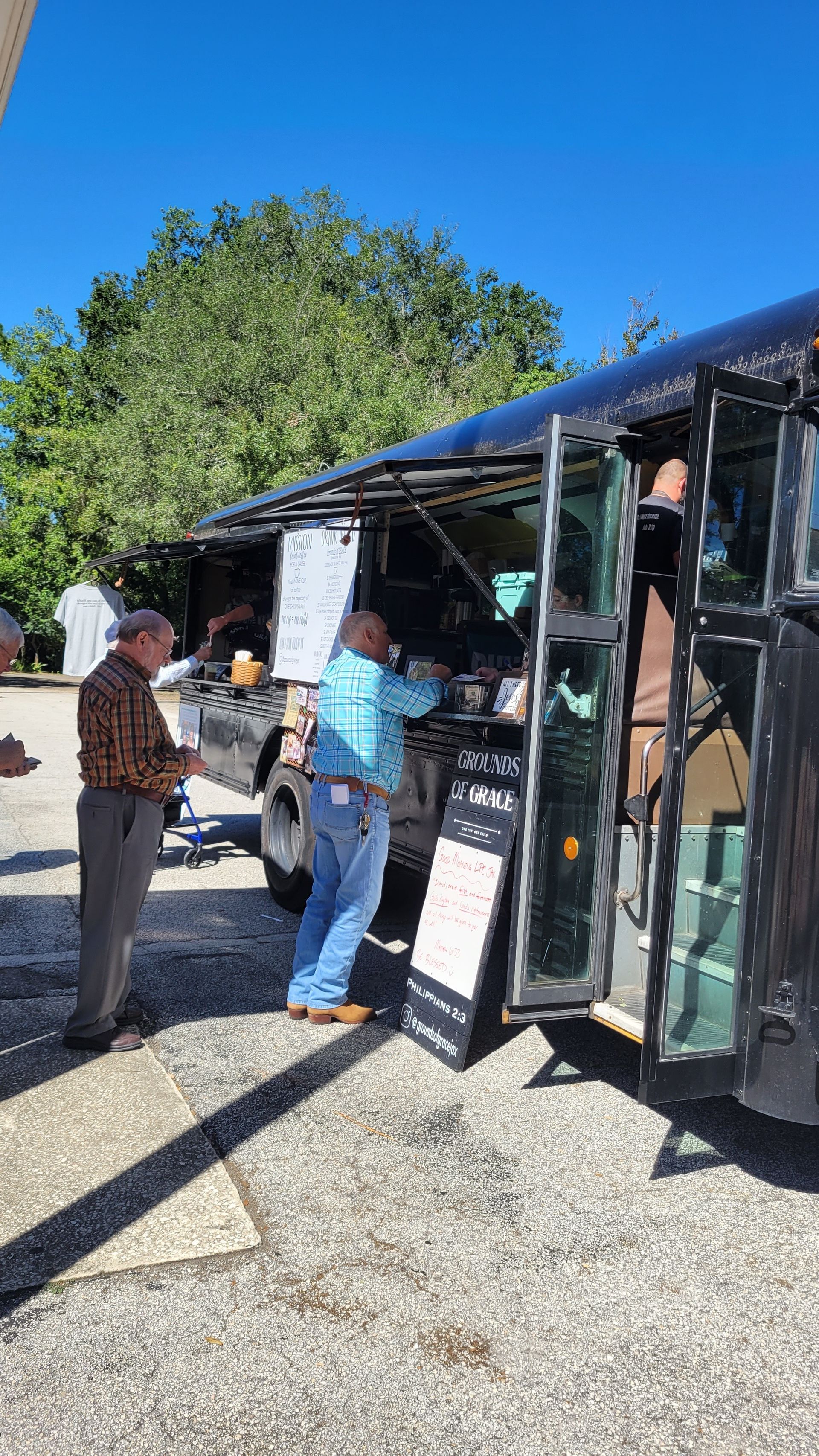 People at a food truck, ordering food. Blue sky and trees in the background.