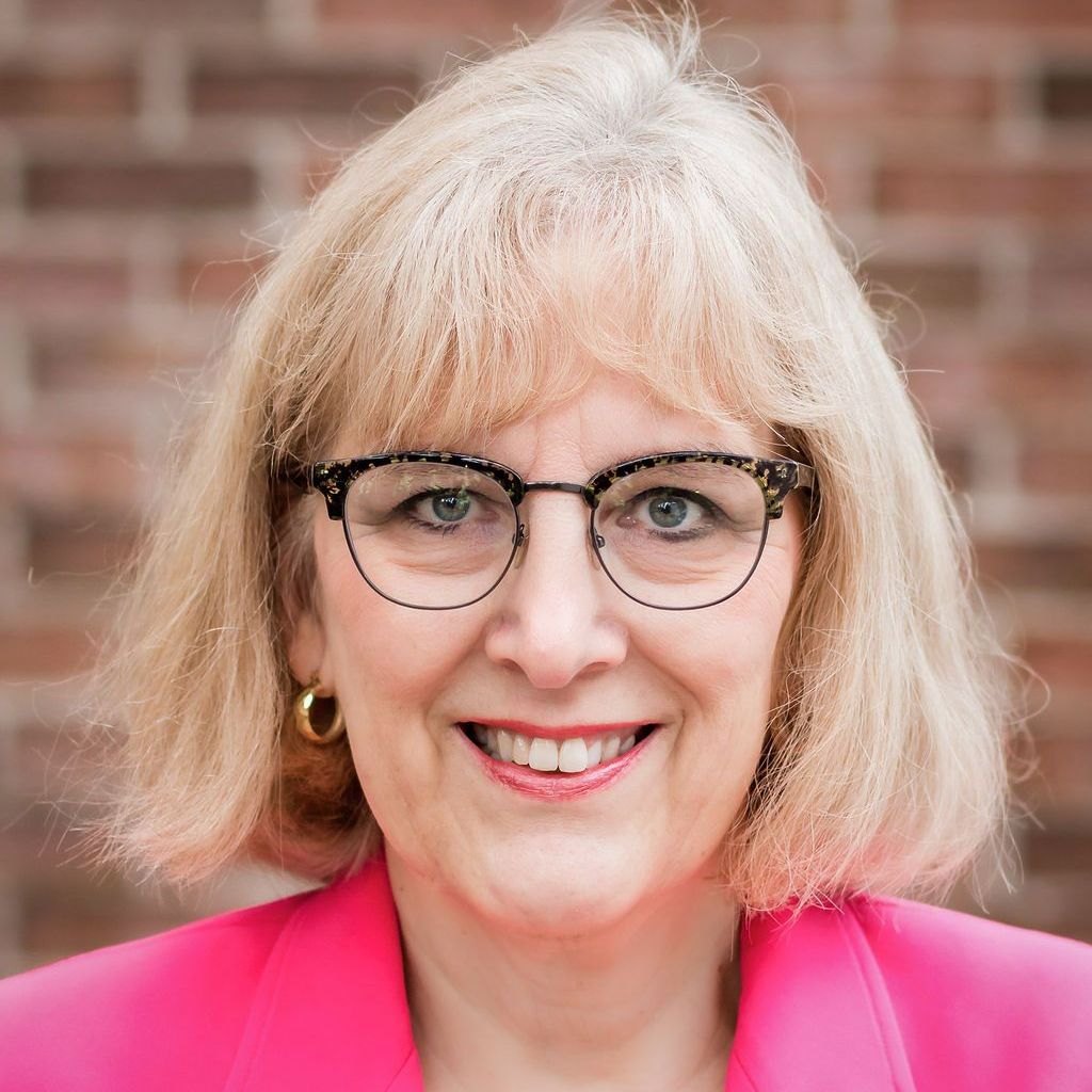 Woman with glasses and blonde hair smiling in front of a brick wall, wearing a pink blazer.
