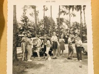 Group of people outdoors near cars and trees, holding food or drinks. Dated August 1962.