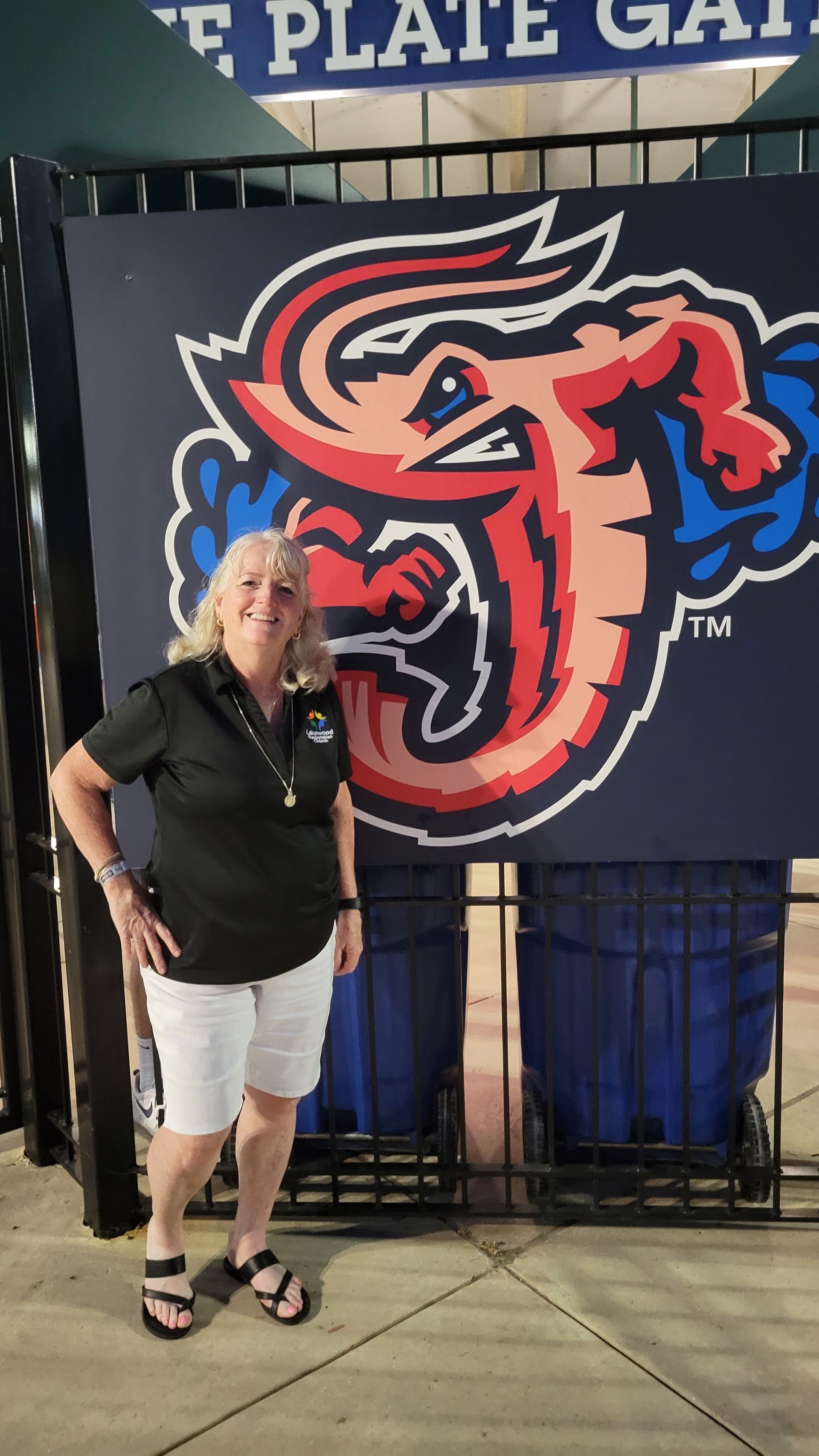 Woman stands by a large, colorful team logo. Logo is a grinning shrimp. Setting appears to be a stadium entrance.