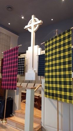 A large white Celtic cross stands behind several tartan banners in yellow, pink, and blue, set inside a church chancel.