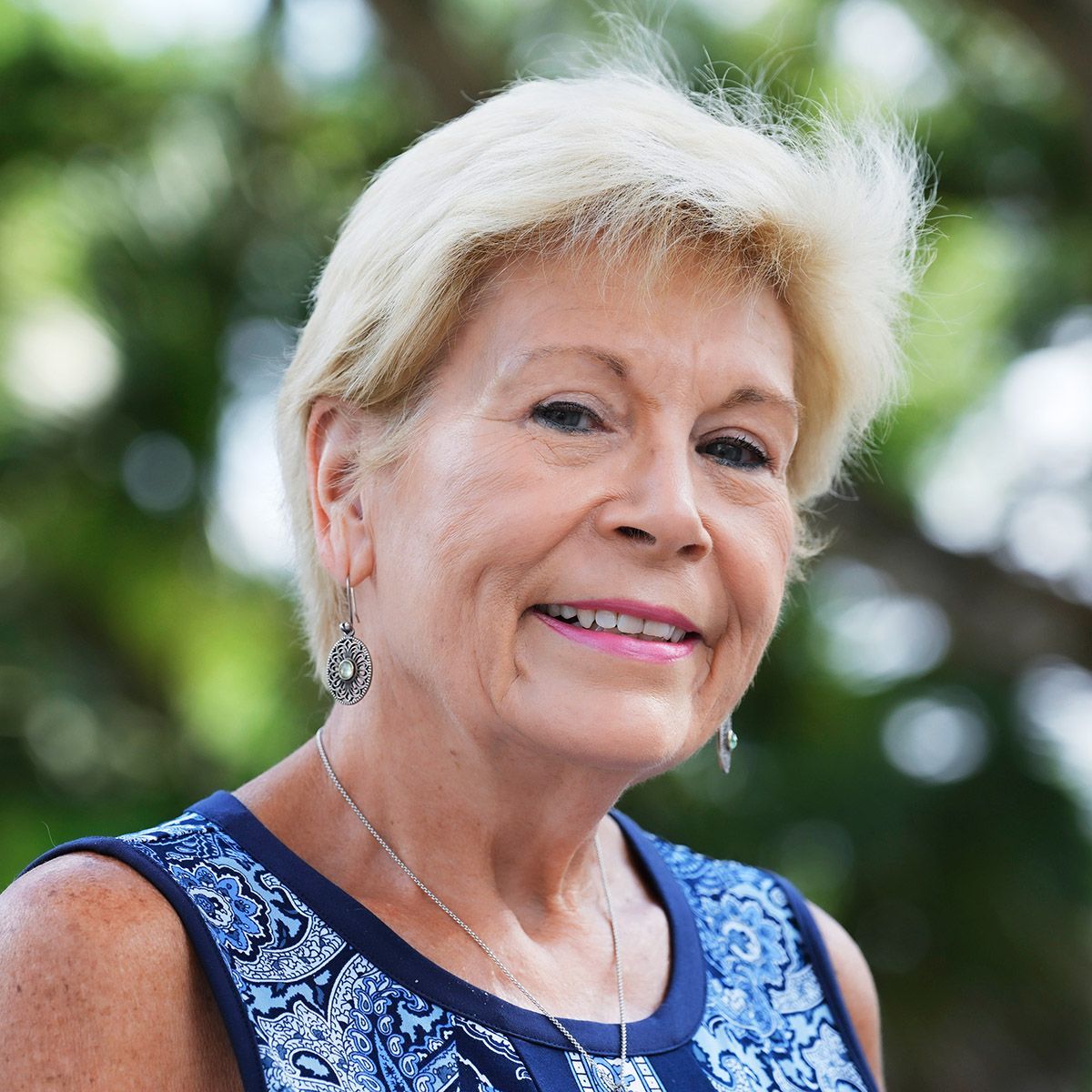 Woman with short blonde hair smiles outdoors, wearing a blue patterned top and small silver earrings.
