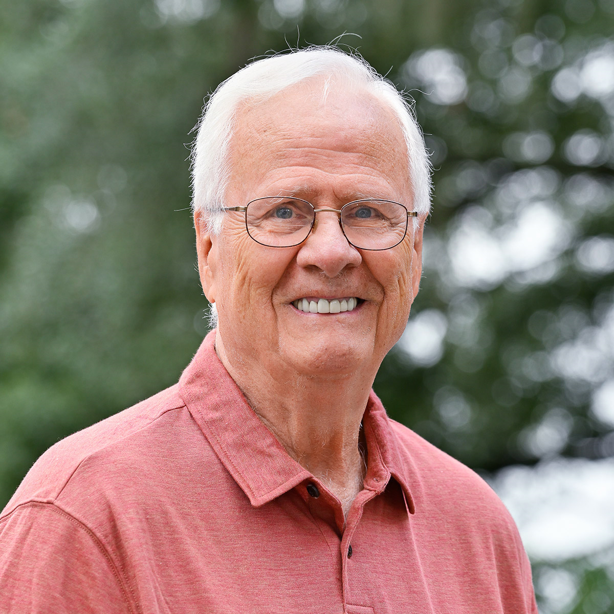 Smiling man with white hair, glasses, and red shirt outdoors.