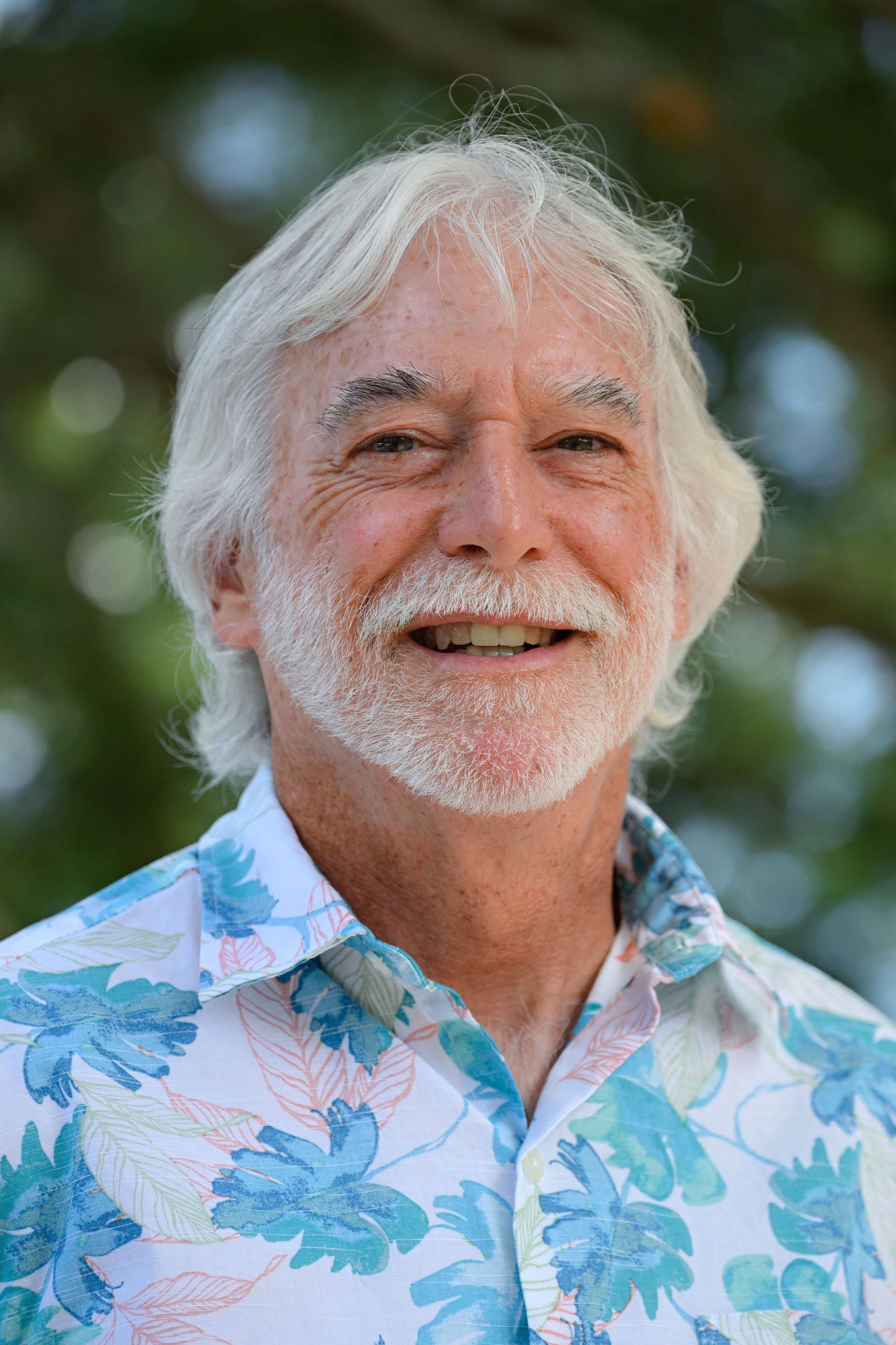 Smiling man with white hair and beard, wearing a floral shirt, outdoors.