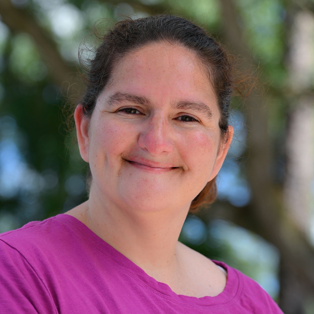 Woman with brown hair smiles, wearing a pink shirt, outdoors with blurred tree background.