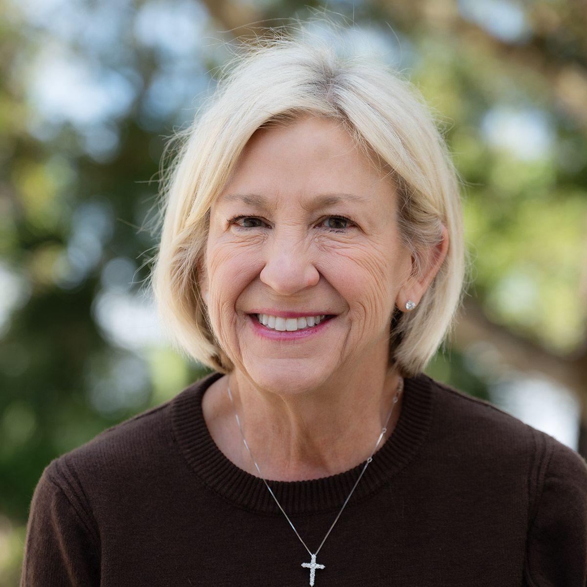 Woman with blonde hair smiling, wearing a brown sweater and a silver cross necklace.