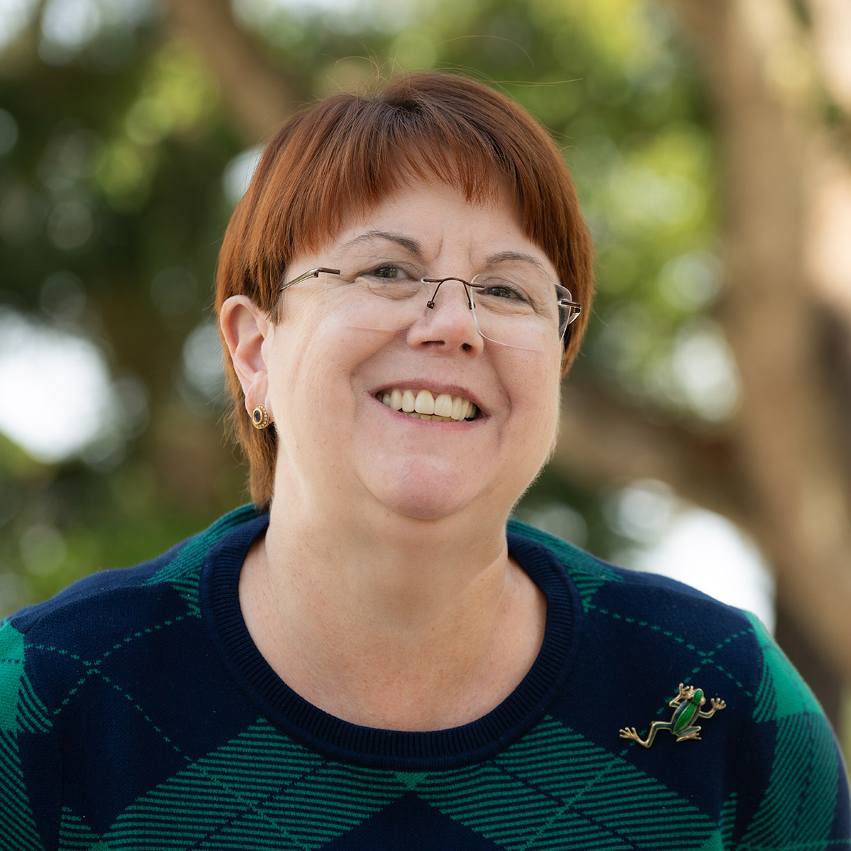 Woman with auburn hair, glasses, smiling, wearing a green and blue plaid sweater with a frog pin; outdoor setting.