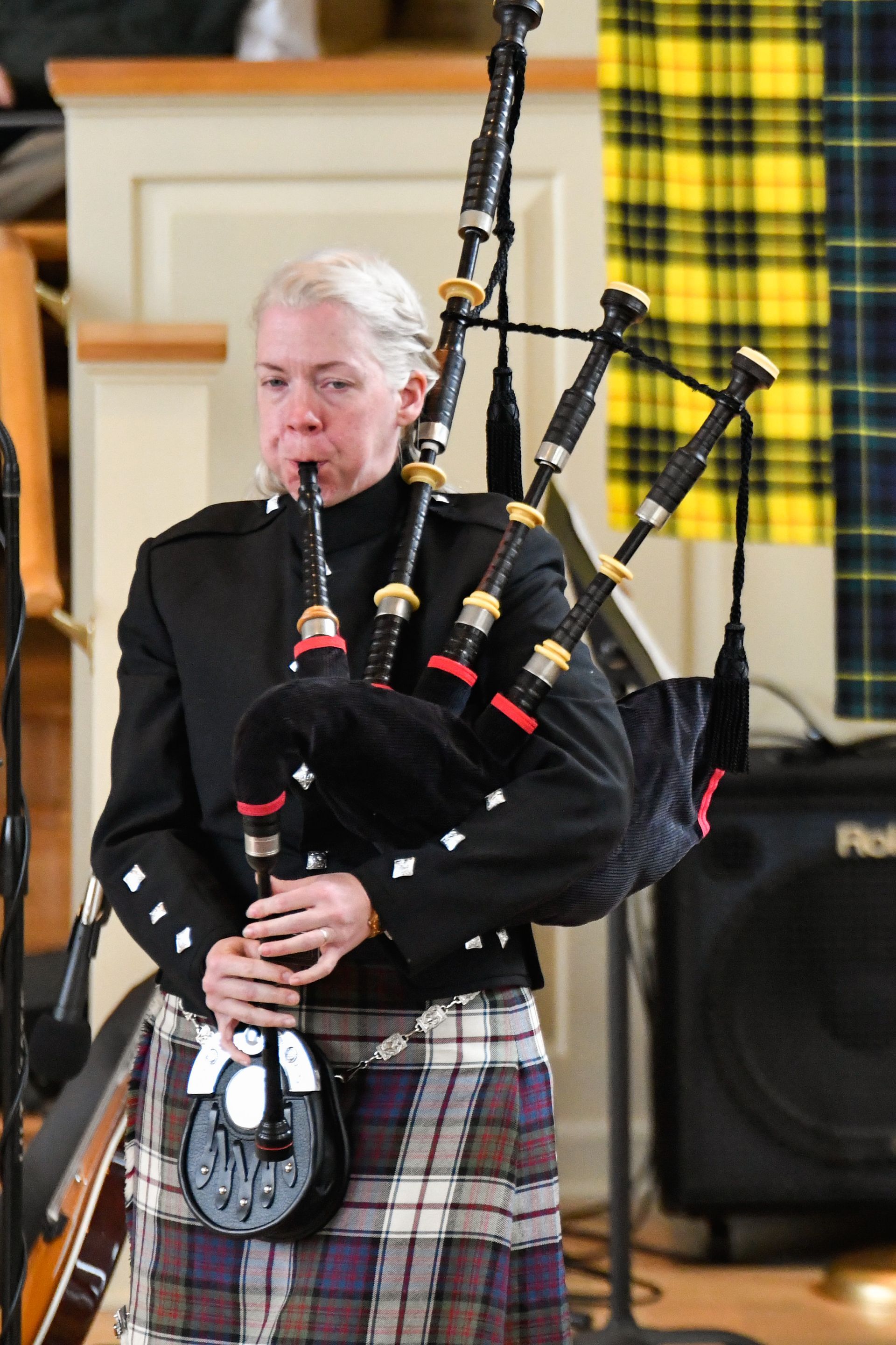 Person playing bagpipes, wearing a kilt and black jacket, indoors near a yellow and black tartan.