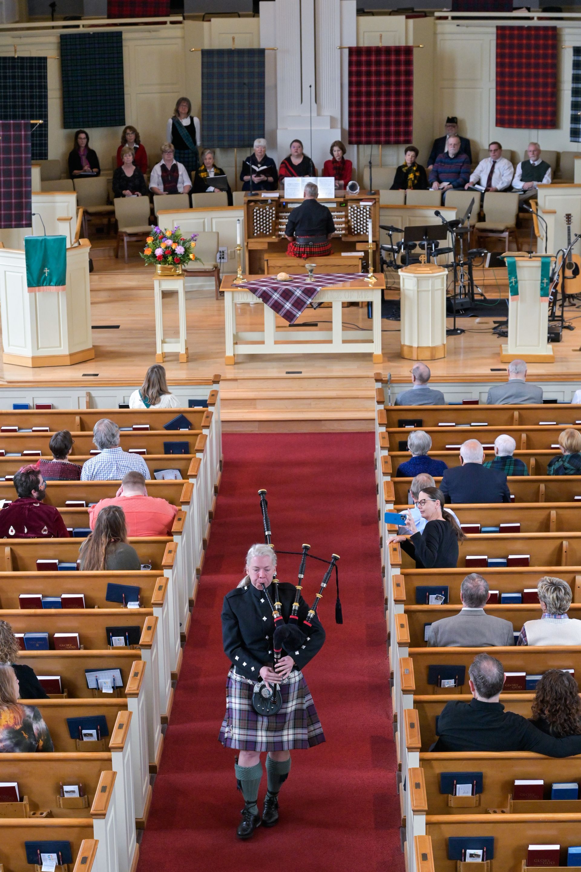 Bagpiper in kilt plays as he walks down church aisle lined with pews. People watch from pews and raised platform.