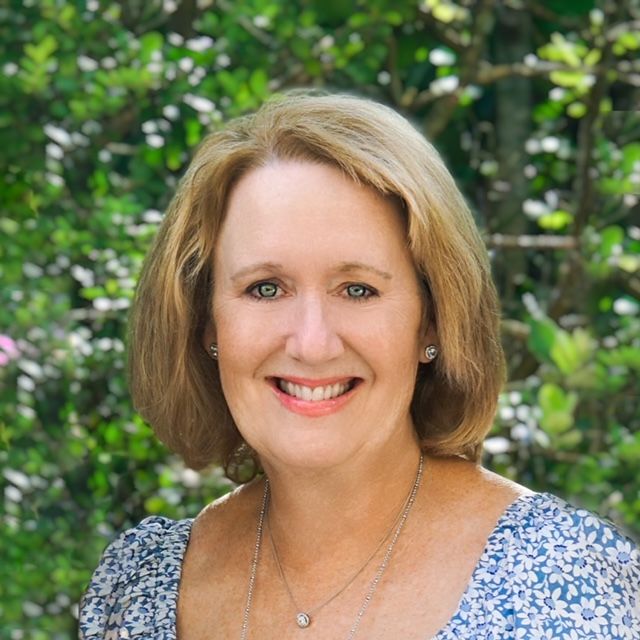 Woman with blonde hair smiles, wearing a floral top, against a green leafy backdrop.