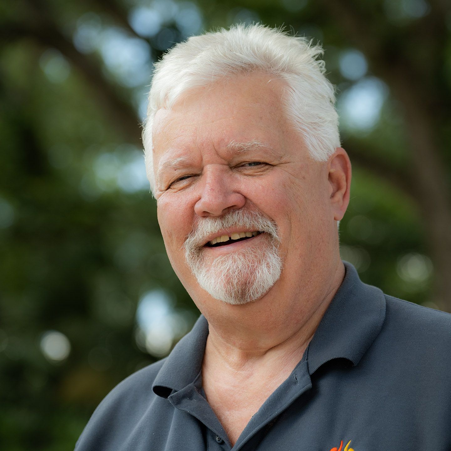 Smiling man with gray hair and goatee wearing a dark blue polo shirt outdoors.