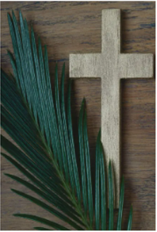 A light-colored wooden cross resting on a dark wooden surface next to a green palm frond.