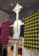 A large white Celtic cross stands behind rows of empty chairs, flanked by several colorful plaid flags on poles.