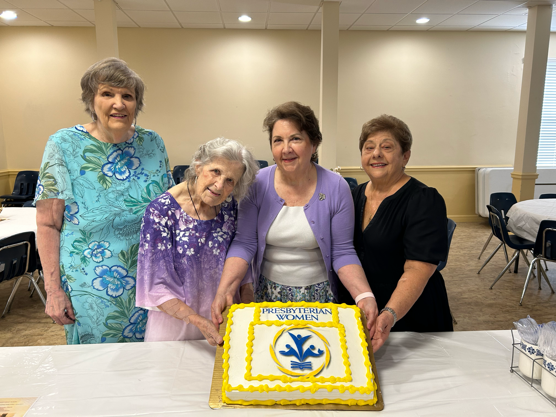 Four women hold a cake decorated with a yellow and blue crest, celebrating at a gathering.